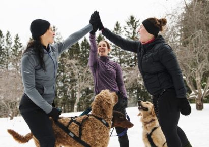 Un groupe de 3 femmes en tenue de sport se tapent dans la main avec leurs chiens tenus en laisse